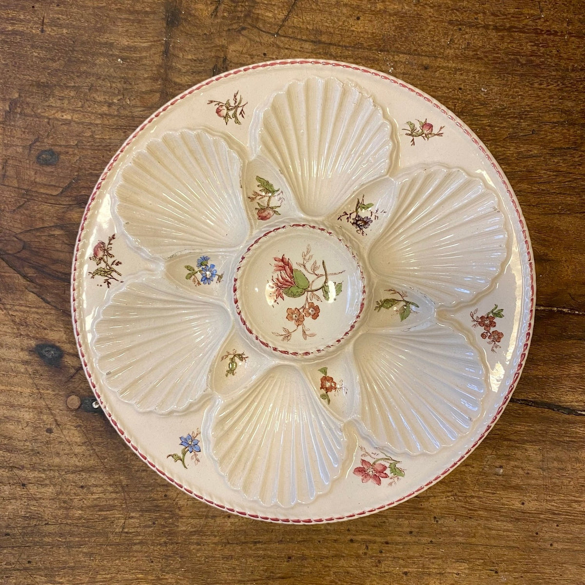 Vintage French oyster plate in off-white with floral sprays and a pink braided rim, seen from above, with six shell-shaped wells around a central compartment