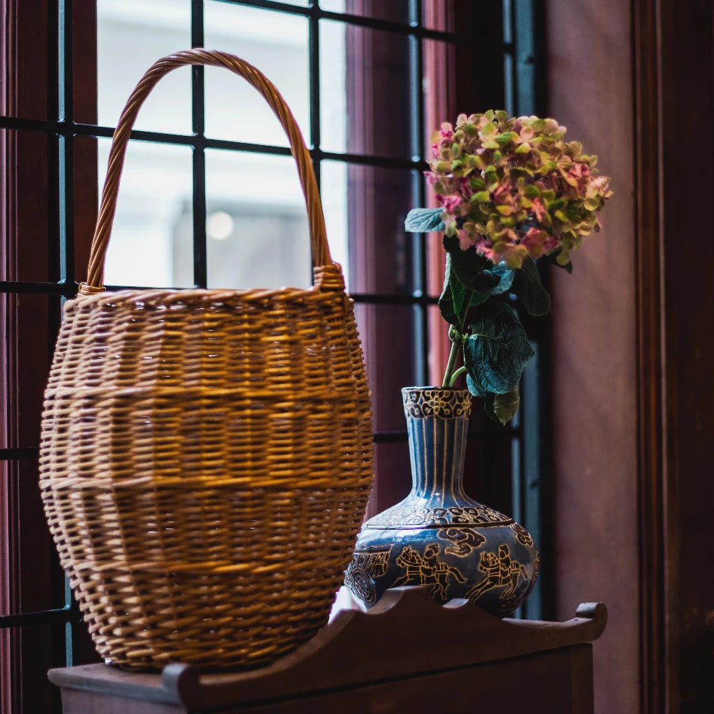 A big colorful vase filled with flowers and a vintage basket place on a wooden cabinet in front of a large window.