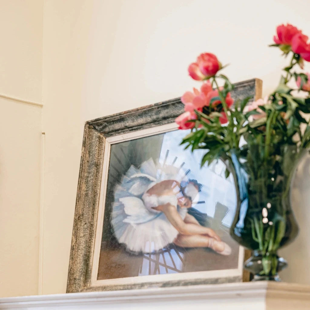 Framed painting of a ballerina resting on a cabinet, with pink flowers in a vase nearby.