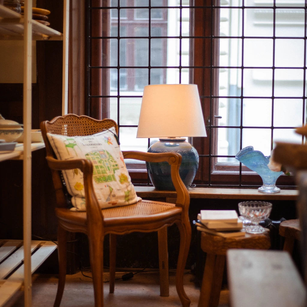 Cozy corner with a vintage chair, decorative pillow, and blue lamp by a window with grid framing.