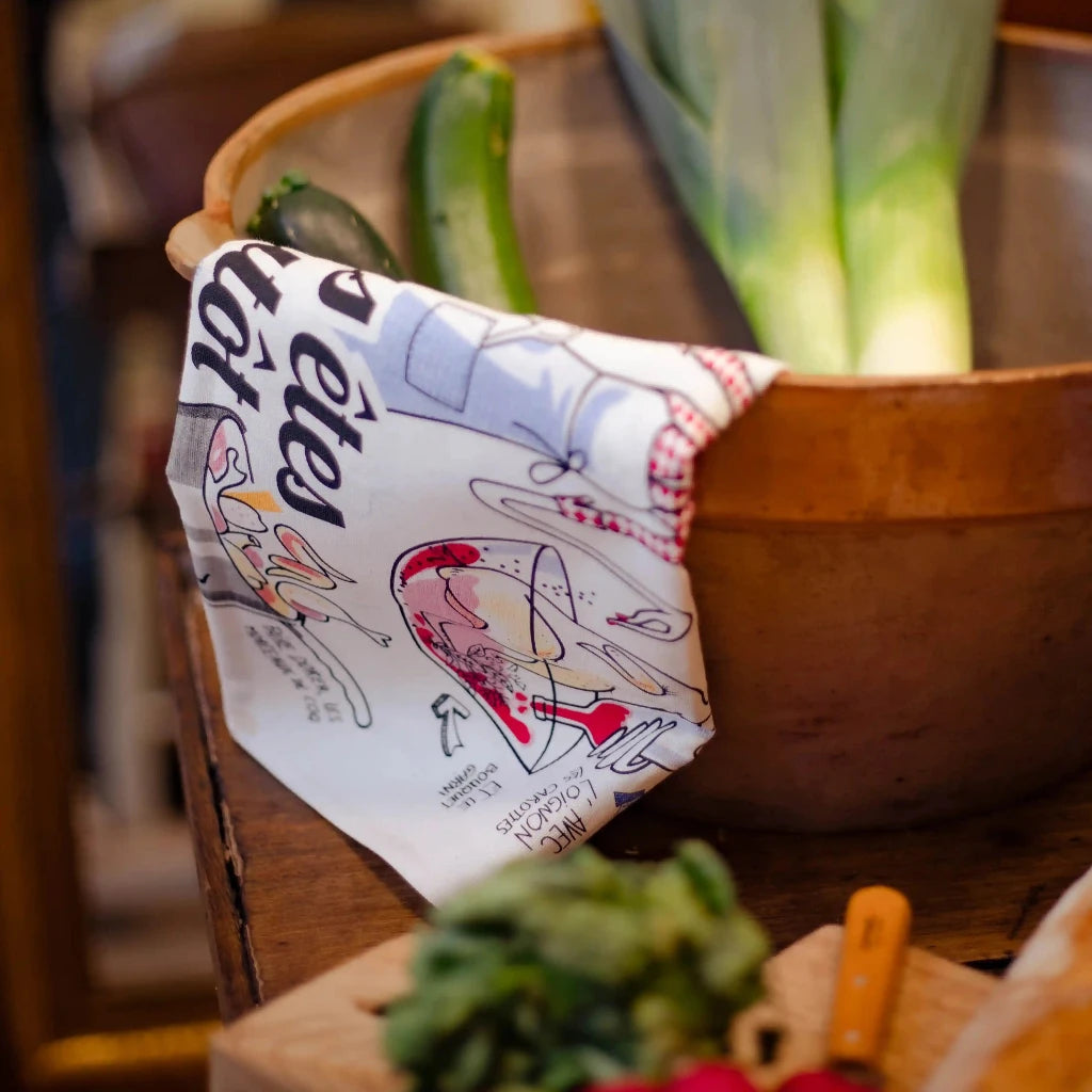 Wooden bowl with fresh vegetables and a colorful kitchen towel with French text and illustrations.