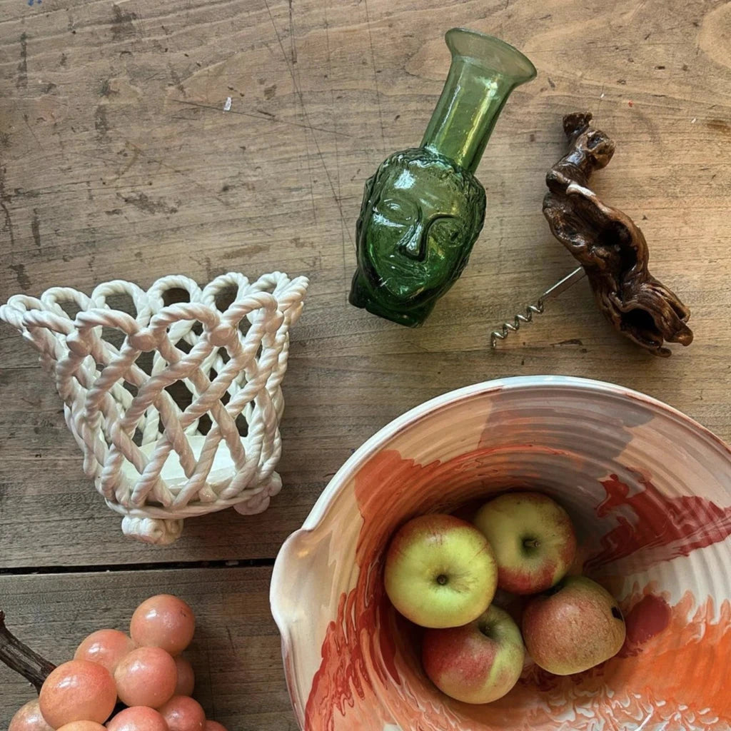 Decorative setup with a ceramic bowl, apples, glass grapes, woven basket, and a green glass vase.